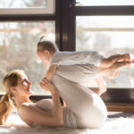 Mama cvičí na podložke doma spolu s malým dieťaťom, ktoré sa hrá vedľa nej.