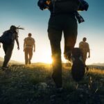 Skupina turistov kráča pri západe slnka po horskom chodníku v letnej obuvi a s batohmi.