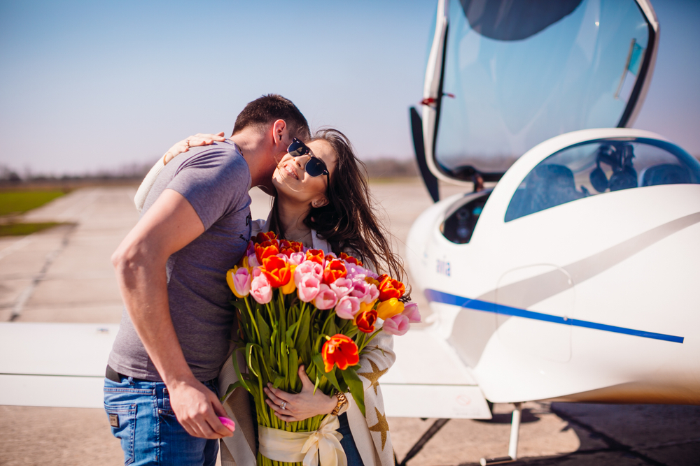 Zamilovaný pár objímajúci sa pred malým lietadlom, žena drží veľkú kyticu tulipánov a usmieva sa so slnečnými okuliarmi na tvári.